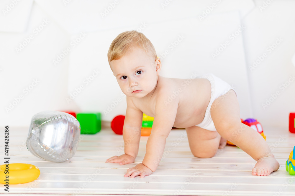 a small baby boy six months old crawls through in a light white nursery in diapers among toys