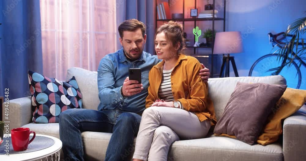 Caucasian young married couple wife and husband resting at home sitting on sofa in modern living room in evening browsing online on smartphone and talking spending time together, marriage concept Caucasian young married couple wife and husband resting at home sitting on sofa in modern living room in evening browsing online on smartphone and talking spending time together, marriage concept