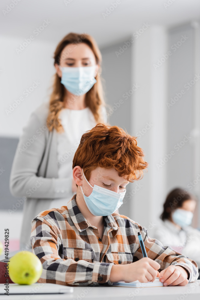 redhead schoolboy in medical mask writing in notebook near teacher on blurred background