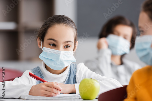 girl in medical mask holding pen near apple and classmate on blurred foreground