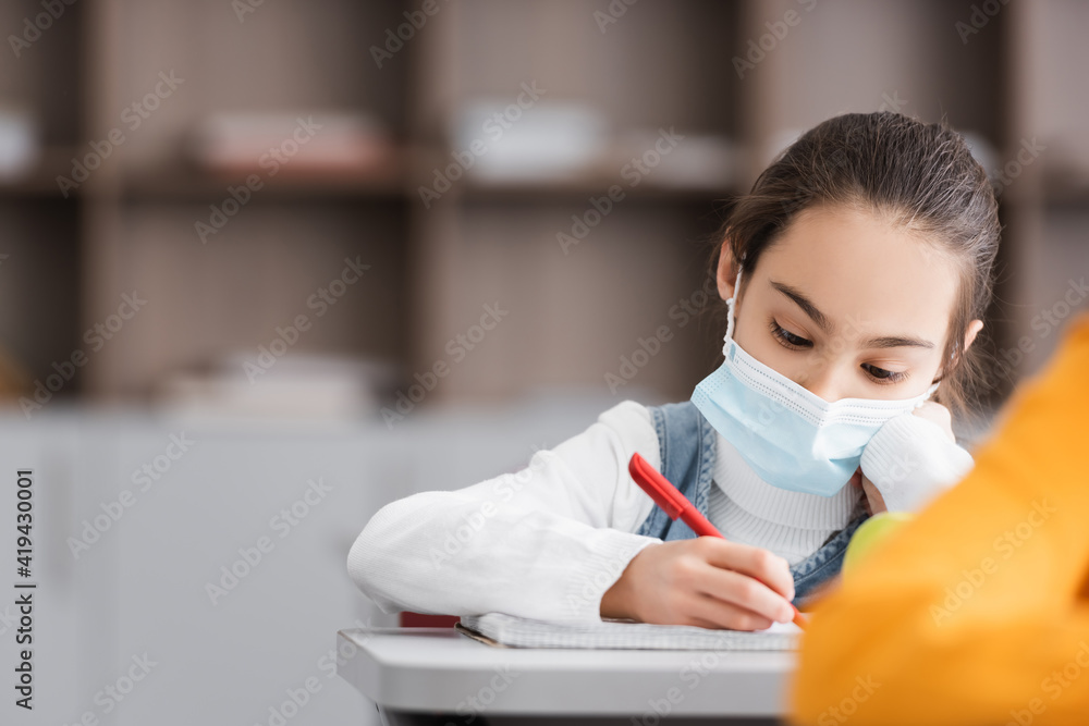 schoolgirl in medical mask writing in notebook on blurred foreground