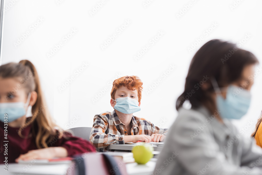 Schoolkid in medical mask looking at camera near classmates on blurred foreground Schoolkid in medical mask looking at camera near classmates on blurred foreground