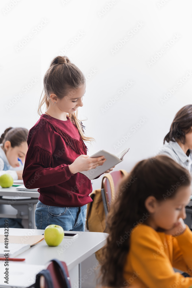 Cheerful schoolgirl reading book near classmates in classroom Cheerful schoolgirl reading book near classmates in classroom