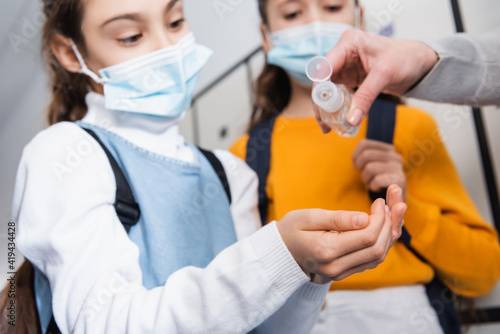 Teacher pouring hand sanitizer on hands of pupil in medical mask on blurred background