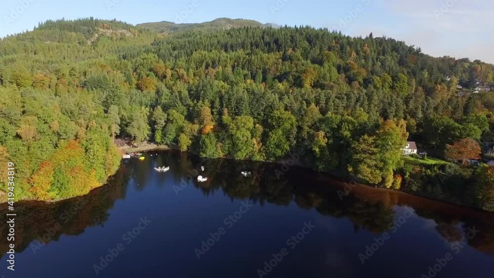 Aerial Forward: Small Boats Float In Still Lake Surrounded By Dense Autumnal Forest And Tall Pines On Hills In Sunshine Aerial Forward: Small Boats Float In Still Lake Surrounded By Dense Autumnal Forest And Tall Pines On Hills In Sunshine