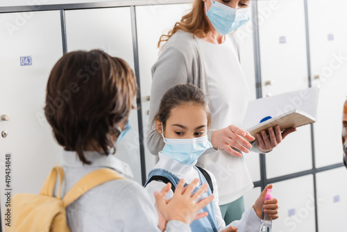 Schoolgirl holding disinfector near friend and teacher with notebook on blurred background