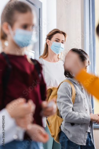 Teacher in medical mask standing near pupils on blurred foreground in hall
