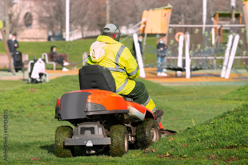 gardener with lawn mower vehicle cutting green grass on city garden