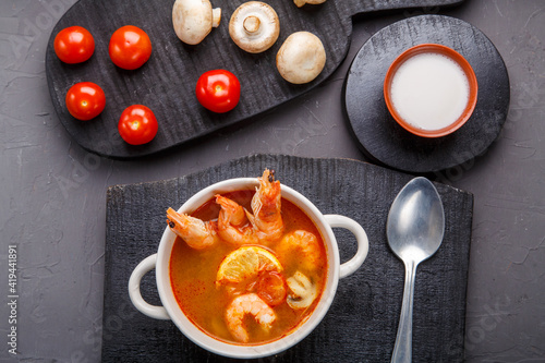 Tom yam soup with shrimps in a tureen on a concrete background next to a bowl with coconut milk tomatoes and mushrooms on a wooden stand.