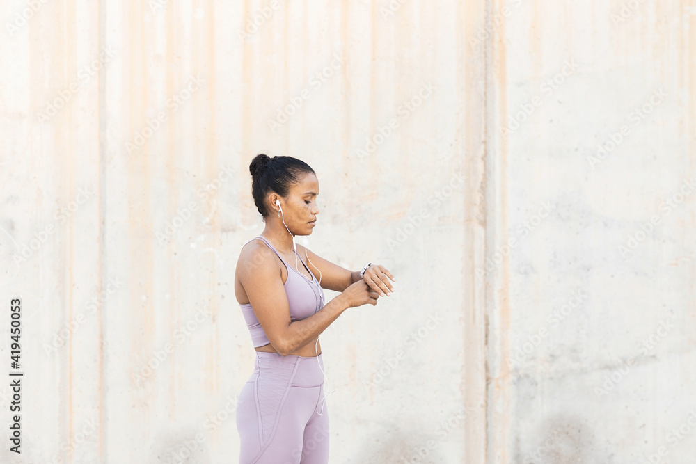 Side view of young ethnic sportswoman checking time while listening to music from earphones against rough wall Side view of young ethnic sportswoman checking time while listening to music from earphones against rough wall