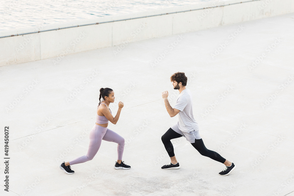 Side view of young determined diverse athletes in sportswear exercising and looking at each other against sea Side view of young determined diverse athletes in sportswear exercising and looking at each other against sea