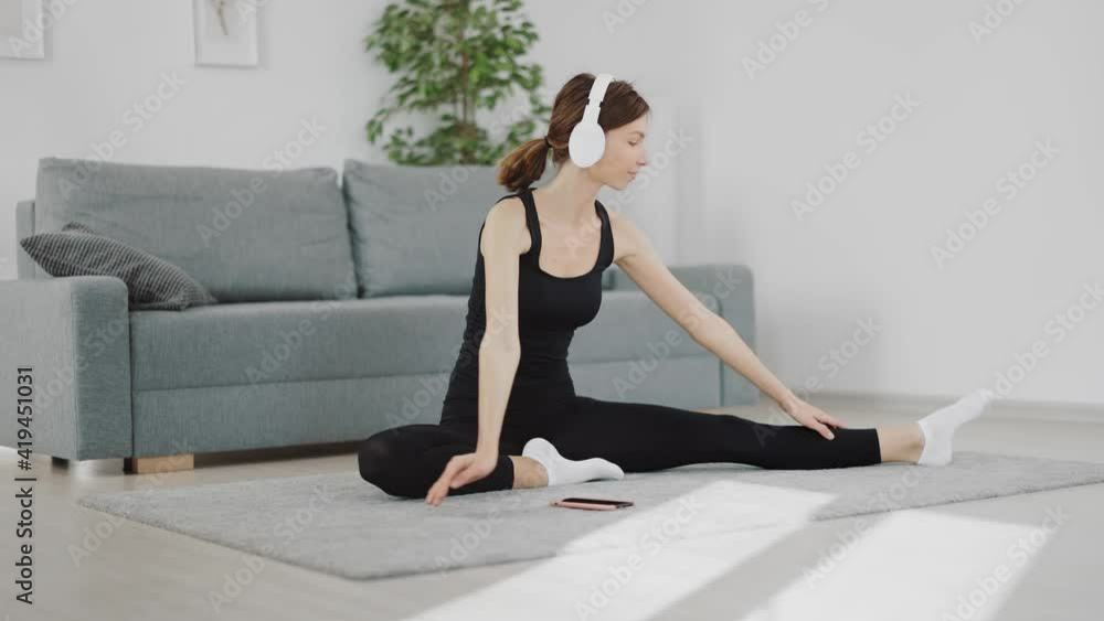 Pleasant sporty woman using wireless headphones and smartphone for domestic workout. Young brunette in black activewear enjoying morning training at home. Pleasant sporty woman using wireless headphones and smartphone for domestic workout. Young brunette in black activewear enjoying morning training at home.