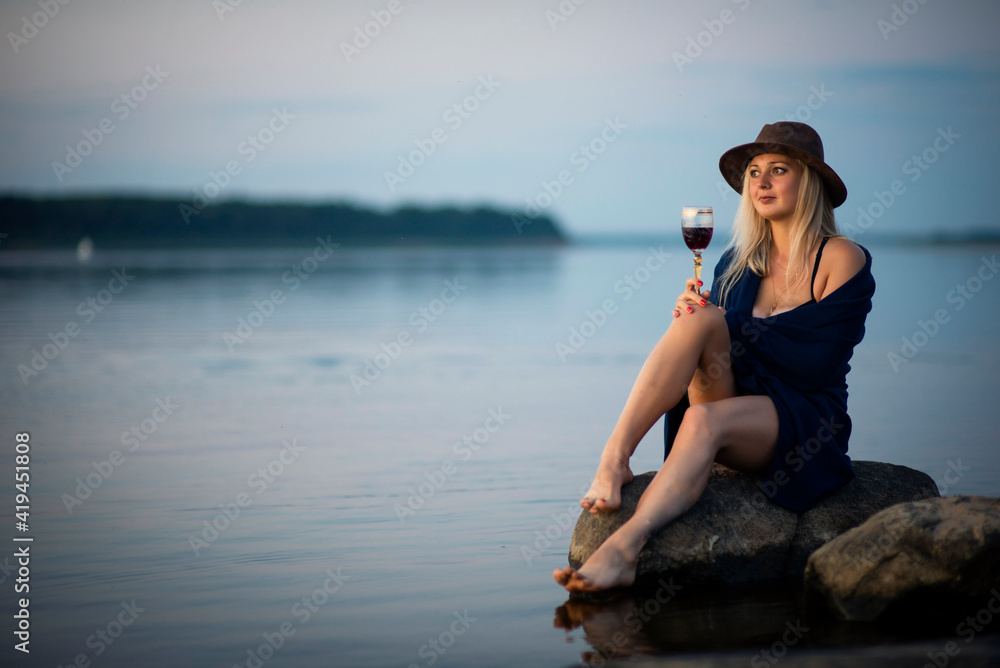 blonde girl in a hat with a glass of wine sitting on the riverbank, selective focus blonde girl in a hat with a glass of wine sitting on the riverbank, selective focus
