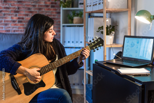 Side view of female playing acoustic guitar while composing music near table with laptop in room with brick wall during remote work