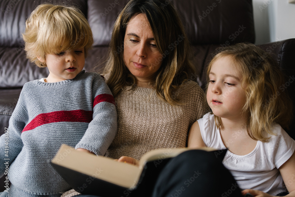 Young woman reading interesting fairytale in book while sitting on floor with little son and daughter at home Young woman reading interesting fairytale in book while sitting on floor with little son and daughter at home
