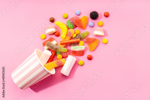 Top view of assorted multicolored sweet jelly candies and marshmallows scattered near disposable striped cup on pink background