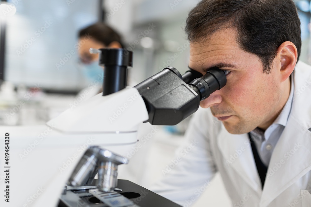 Concentrated male scientist using microscope and analyzing chemical samples while female colleague working on laptop in modern laboratory Concentrated male scientist using microscope and analyzing chemical samples while female colleague working on laptop in modern laboratory