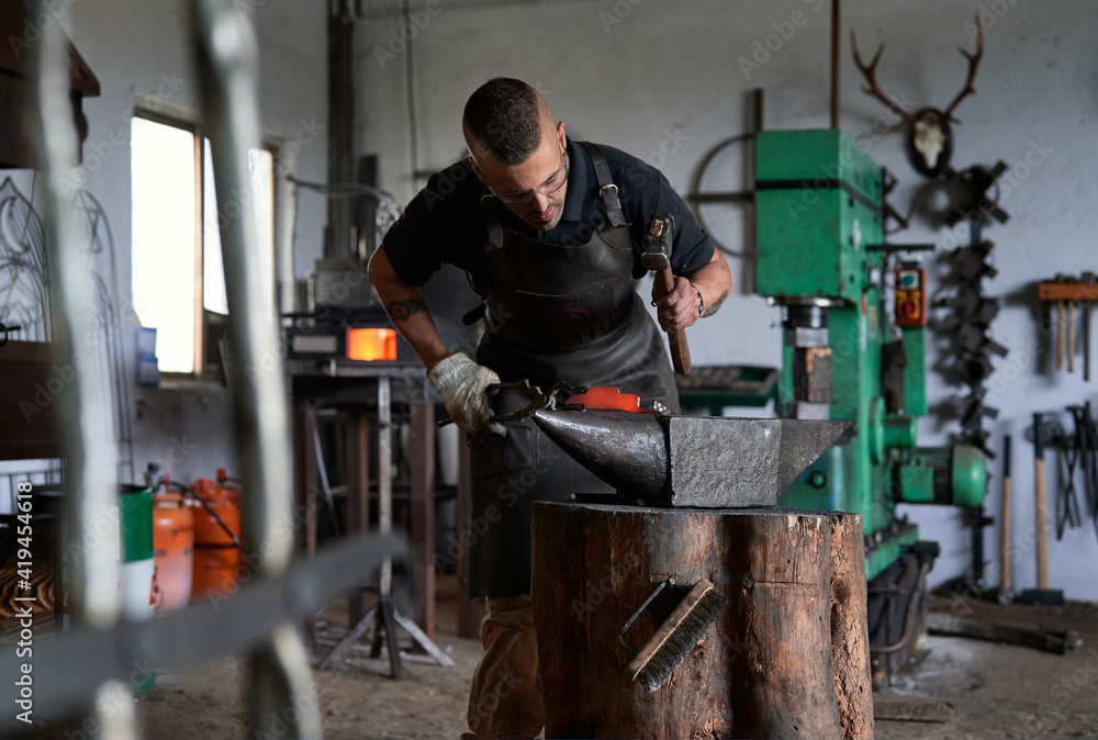 Front view of concentrated young male blacksmith in casual clothes and apron working near anvil with hammer in hand Front view of concentrated young male blacksmith in casual clothes and apron working near anvil with hammer in hand
