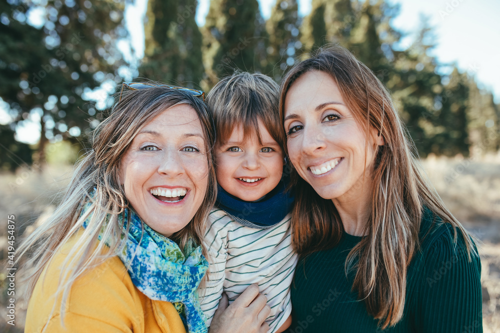Joyful lesbian couple embracing cute little son and looking at camera with friendly smiles while spending sunny day in peaceful nature Joyful lesbian couple embracing cute little son and looking at camera with friendly smiles while spending sunny day in peaceful nature