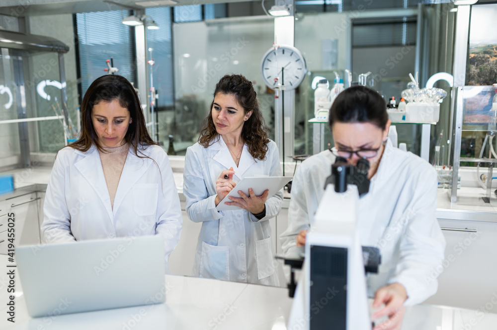 Group of professional female scientists using microscope and laptop while conducting research together in modern lab Group of professional female scientists using microscope and laptop while conducting research together in modern lab