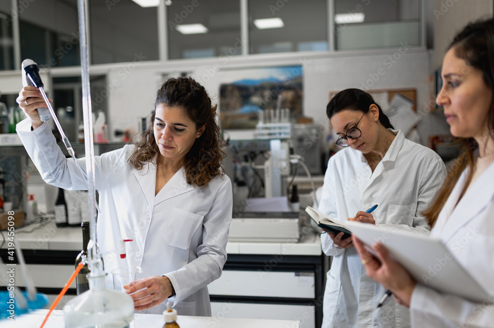 Group of female scientists in white coats examining results of chemical reaction and writing notes in documents while developing new medication in modern lab Group of female scientists in white coats examining results of chemical reaction and writing notes in documents while developing new medication in modern lab