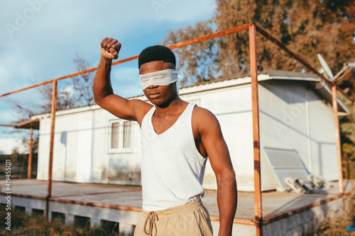 Anonymous masculine ethnic male protester in undershirt and blindfold standing with raised arm in countryside photo
