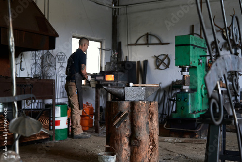 Back view of serious tattooed young male forger in apron heating metal in furnace while working in grungy smithy