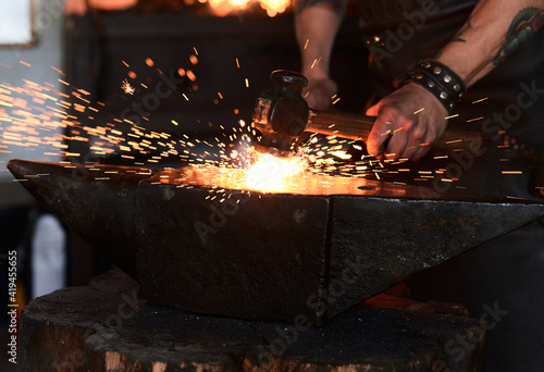 Side view of young brutal bearded craftsman in apron and goggles heating and striking iron on anvil with hammer during work in traditional smithy