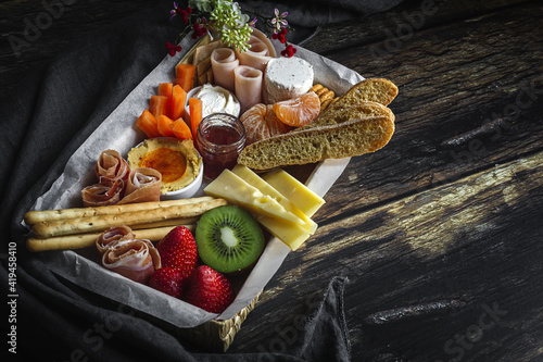 From above brunch box with assorted sliced meats various types of cheese and crispbreads arranged near ripe cup kiwi sweet strawberries and peeled mandarin near jam in glass jar on wooden table near napkin photo