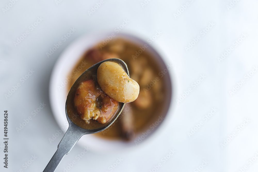 Top view spoon with saucy white beans over bowl with delicious dish with langoustines served on white table Top view spoon with saucy white beans over bowl with delicious dish with langoustines served on white table