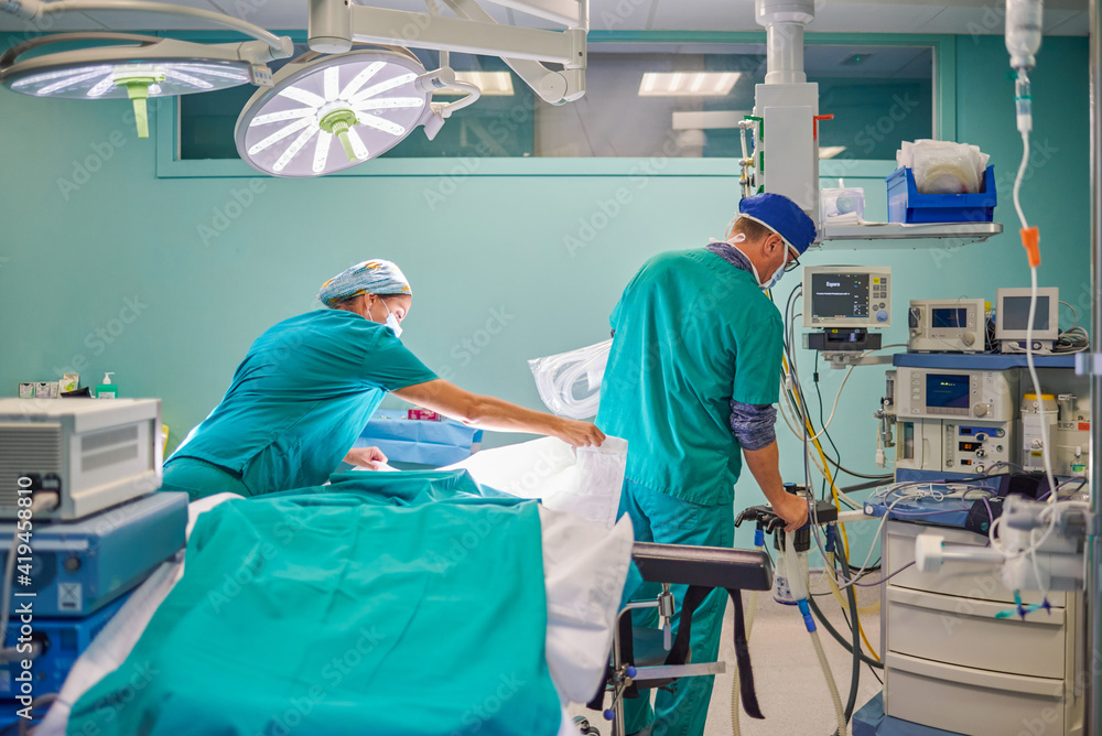 Back view of unrecognizable male and female medical assistants in uniforms preparing couch and devices in operating room for surgery in modern hospital Back view of unrecognizable male and female medical assistants in uniforms preparing couch and devices in operating room for surgery in modern hospital