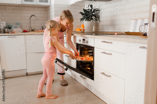 Mom and daughter cook in the kitchen. A young woman cooks with her child in the kitchen at home. Girls in pink clothes cook pizza. Happy and cheerful mom and daughter