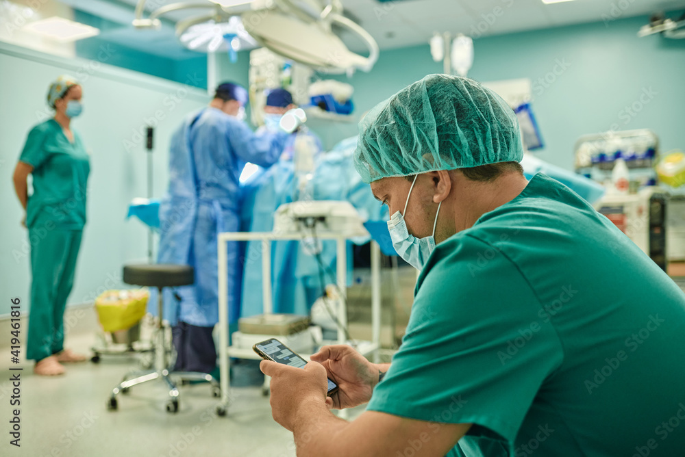 Side view of anonymous concentrated young male assistant in medical uniform and mask messaging on mobile phone while team of professional surgeons performing operation in modern clinic Side view of anonymous concentrated young male assistant in medical uniform and mask messaging on mobile phone while team of professional surgeons performing operation in modern clinic