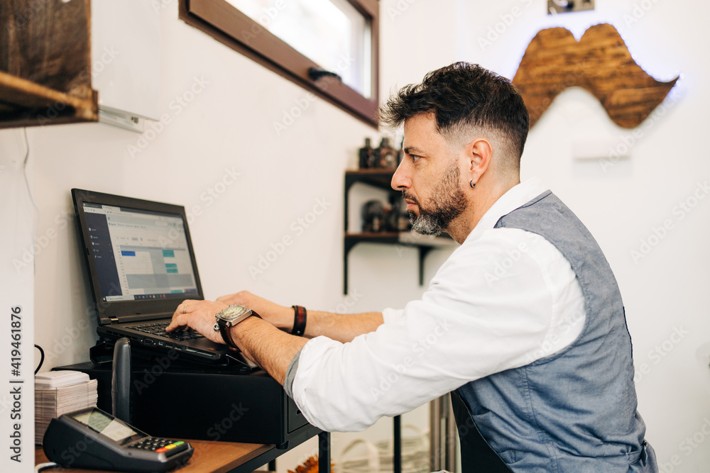 Side view of focused male barber working on netbook with open program on screen in beauty salon near cash register till and card reader Side view of focused male barber working on netbook with open program on screen in beauty salon near cash register till and card reader