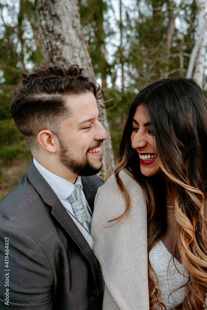 Side view of cheerful young bearded groom in suit laughing and cuddling elegant ethnic bride in white dress while standing together on wooden bridge near aged house in autumn forest Side view of cheerful young bearded groom in suit laughing and cuddling elegant ethnic bride in white dress while standing together on wooden bridge near aged house in autumn forest