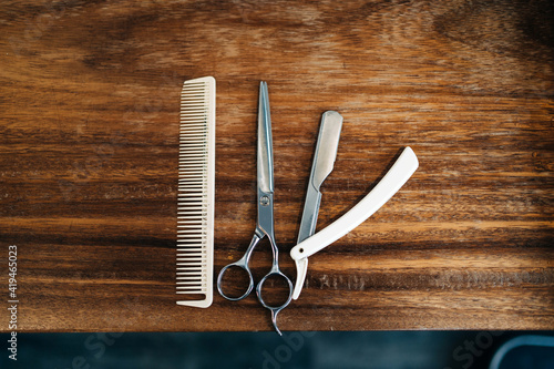 Top view of scissor and comb near straight razor with sharp metal blades on wooden table in hairdressing salon