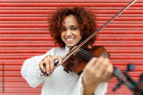 Happy beautiful professional female musician in white sweater playing acoustic violin and looking at camera with toothy smile against red wall photo