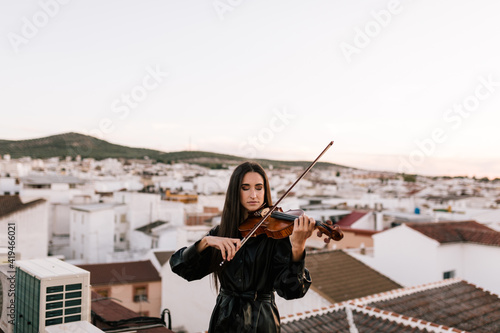 Young beautiful female musician in stylish mini dress holding acoustic violin and standing on rooftop in residential suburb and closed eyes on sunny evening photo
