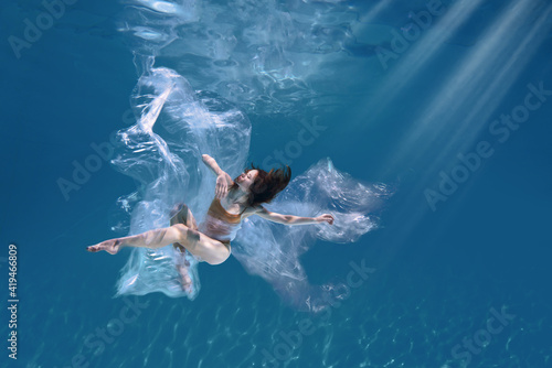 Fashionable and athletic girl free diver alone in the depths of the ocean. Swimmer brunette diving deep in ocean on blue underwater background. Pollution, plastic and ecology concept