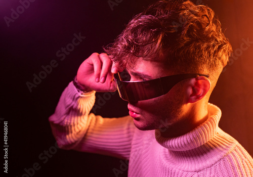 Side view of self assured young male in stylish outfit adjusting futuristic sunglasses while standing in dark studio with neon illumination photo