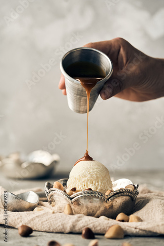 Unrecognizable crop person pouring sweet caramel topping on vanilla ice cream scoop in bowl placed on table with scattered nuts