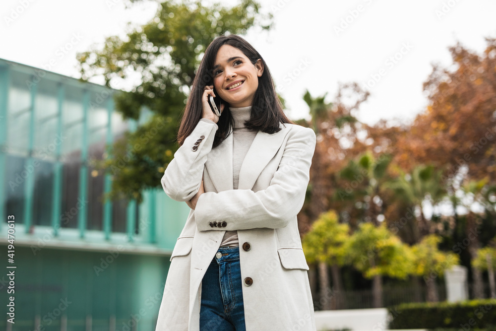 Low angle of glad female entrepreneur talking on smartphone and standing in street while discussing business project and looking away Low angle of glad female entrepreneur talking on smartphone and standing in street while discussing business project and looking away