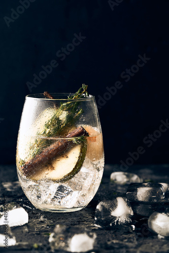 Crystal glass of refreshing infused water with pear slice and ice cubes garnished with herb and cinnamon stick on black background
