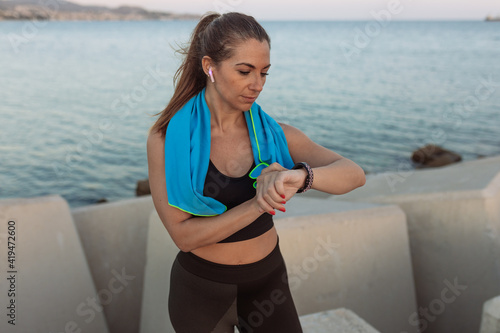 Fit female athlete with wireless earbuds and towel on neck checking results of exercising on fitness tracker while resting on embankment after workout
