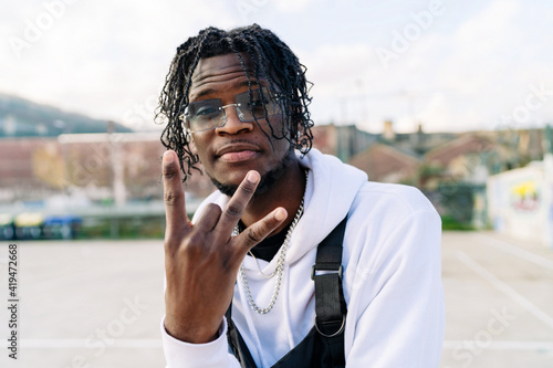 Trendy African American male in sunglasses with Afro braids demonstrating rap gesture while looking at camera in town photo