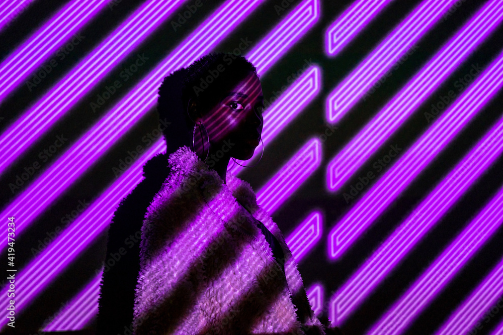 African American woman in stylish fur coat looking at camera while standing under violet neon stripes African American woman in stylish fur coat looking at camera while standing under violet neon stripes