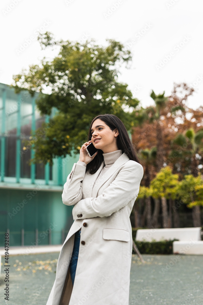Low angle of glad female entrepreneur talking on smartphone and standing in street while discussing business project and looking away Low angle of glad female entrepreneur talking on smartphone and standing in street while discussing business project and looking away