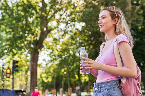 Side view of cheerful young female with backpack and bottle of water standing in green park in sunny day in Madrid