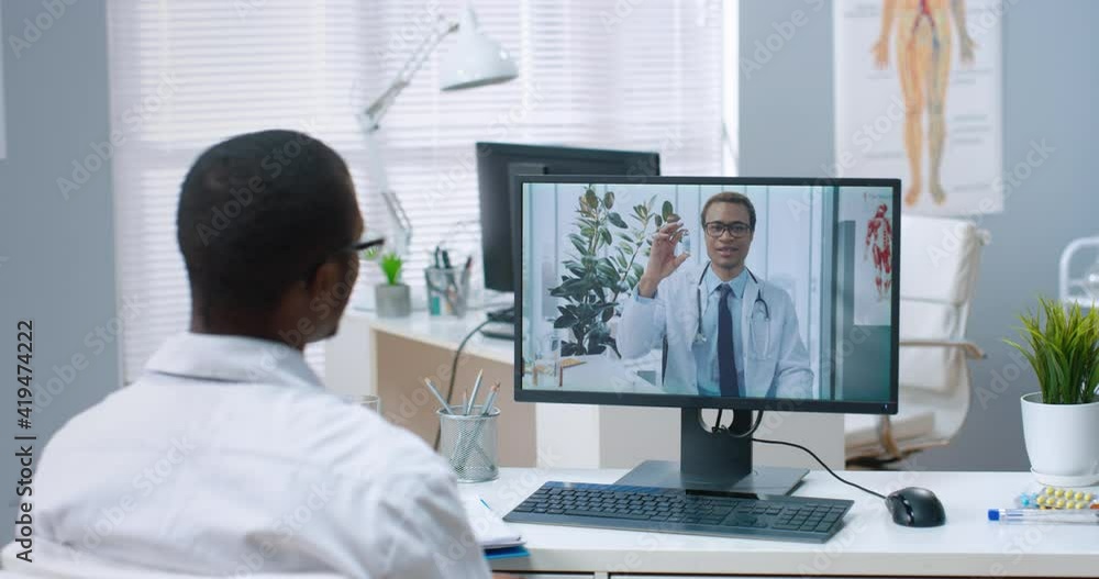 Rear of male African American doctor speaking on video call online consulting with specialist colleague on computer physician gives online consultation on webcam conference about treatment and vaccine Rear of male African American doctor speaking on video call online consulting with specialist colleague on computer physician gives online consultation on webcam conference about treatment and vaccine