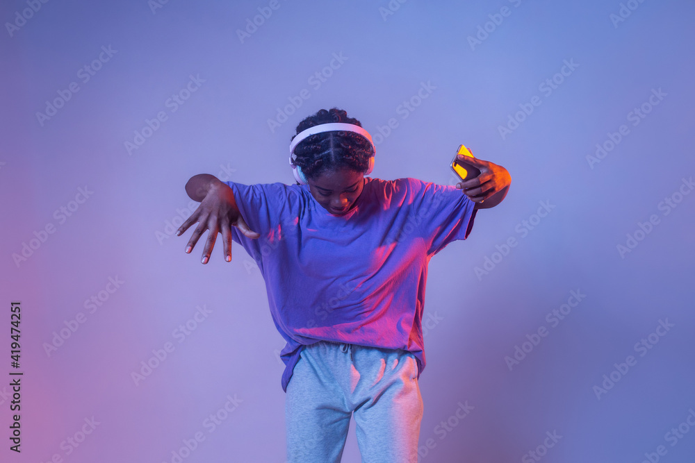Serious African American teenage girl with smartphone in headphones standing in studio with bright glowing neon lights Serious African American teenage girl with smartphone in headphones standing in studio with bright glowing neon lights